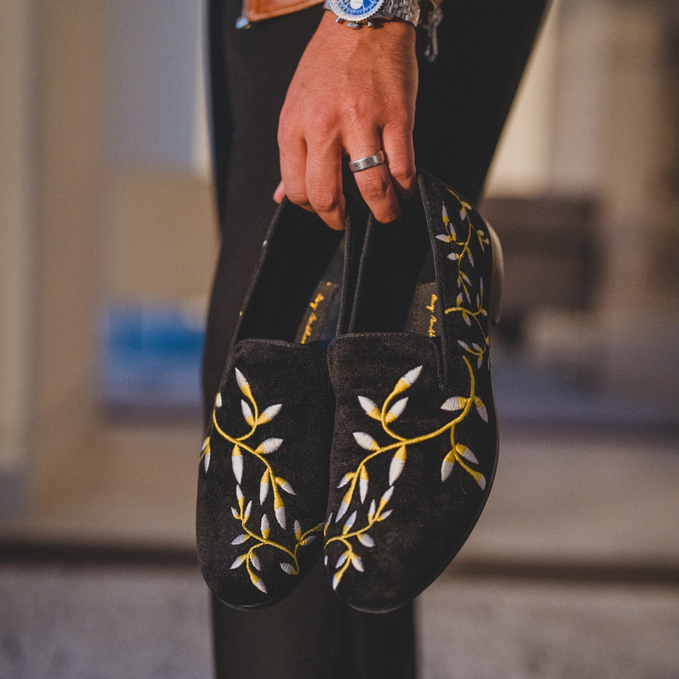 Person holding black slippers with yellow floral embroidery, wearing a brown leather jacket.